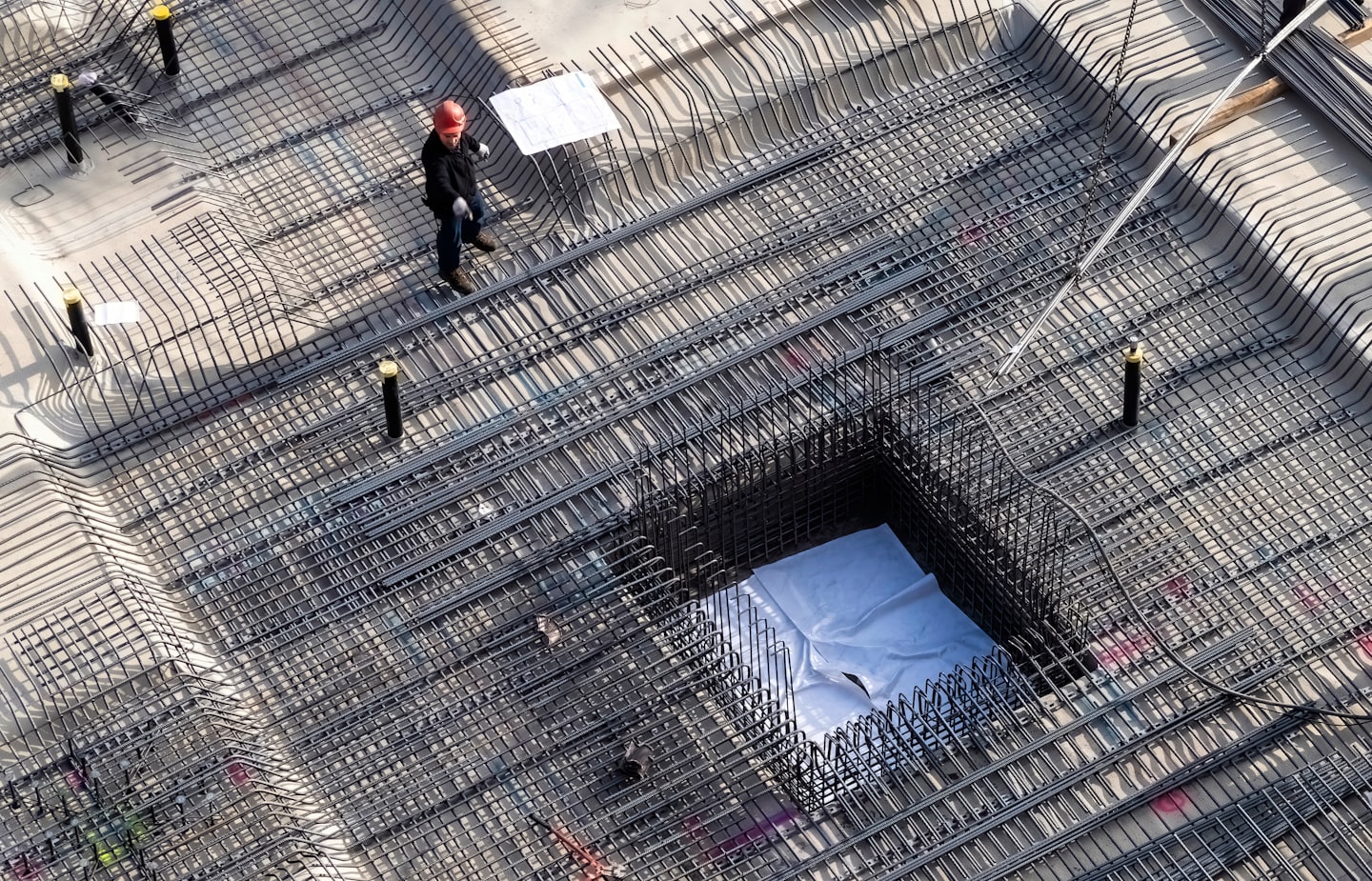 Construction worker inspects blueprints on rebar grid foundation.