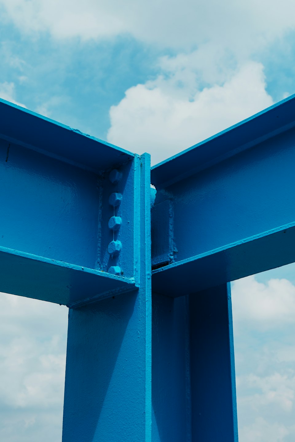 Blue industrial I-beams connected by bolts against a cloudy sky.